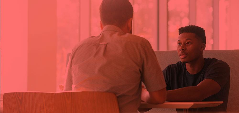 Two people sit across from each other at a table, one seen from behind and the other facing forward, in a booth-style seating area with a red colour overlay.