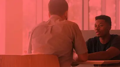 Two people sit across from each other at a table, one seen from behind and the other facing forward, in a booth-style seating area with a red colour overlay.