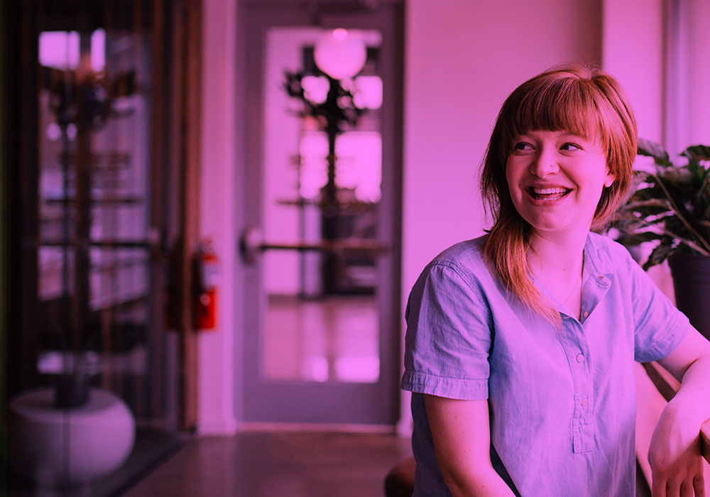 A woman sits indoors, smiling and looking to the side, wearing a short-sleeved shirt. The background shows a doorway, plants, and interior furnishings, with a pink-purple colour overlay.