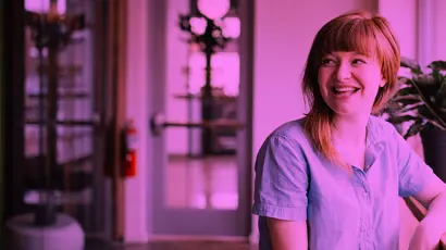 A woman sits indoors, smiling and looking to the side, wearing a short-sleeved shirt. The background shows a doorway, plants, and interior furnishings, with a pink-purple colour overlay.