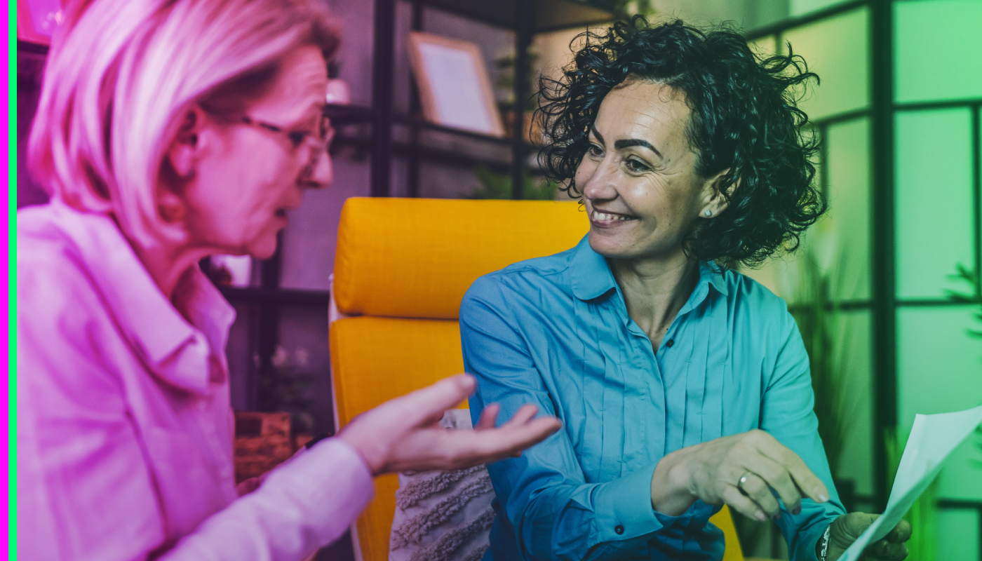Two women sit facing each other indoors, talking and smiling. One woman gestures with her hand while the other holds a sheet of paper.