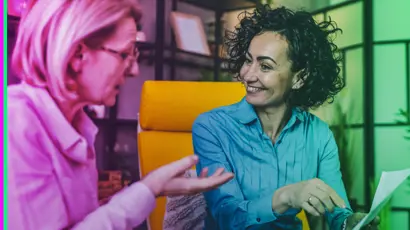 Two women sit facing each other indoors, talking and smiling. One woman gestures with her hand while the other holds a sheet of paper.