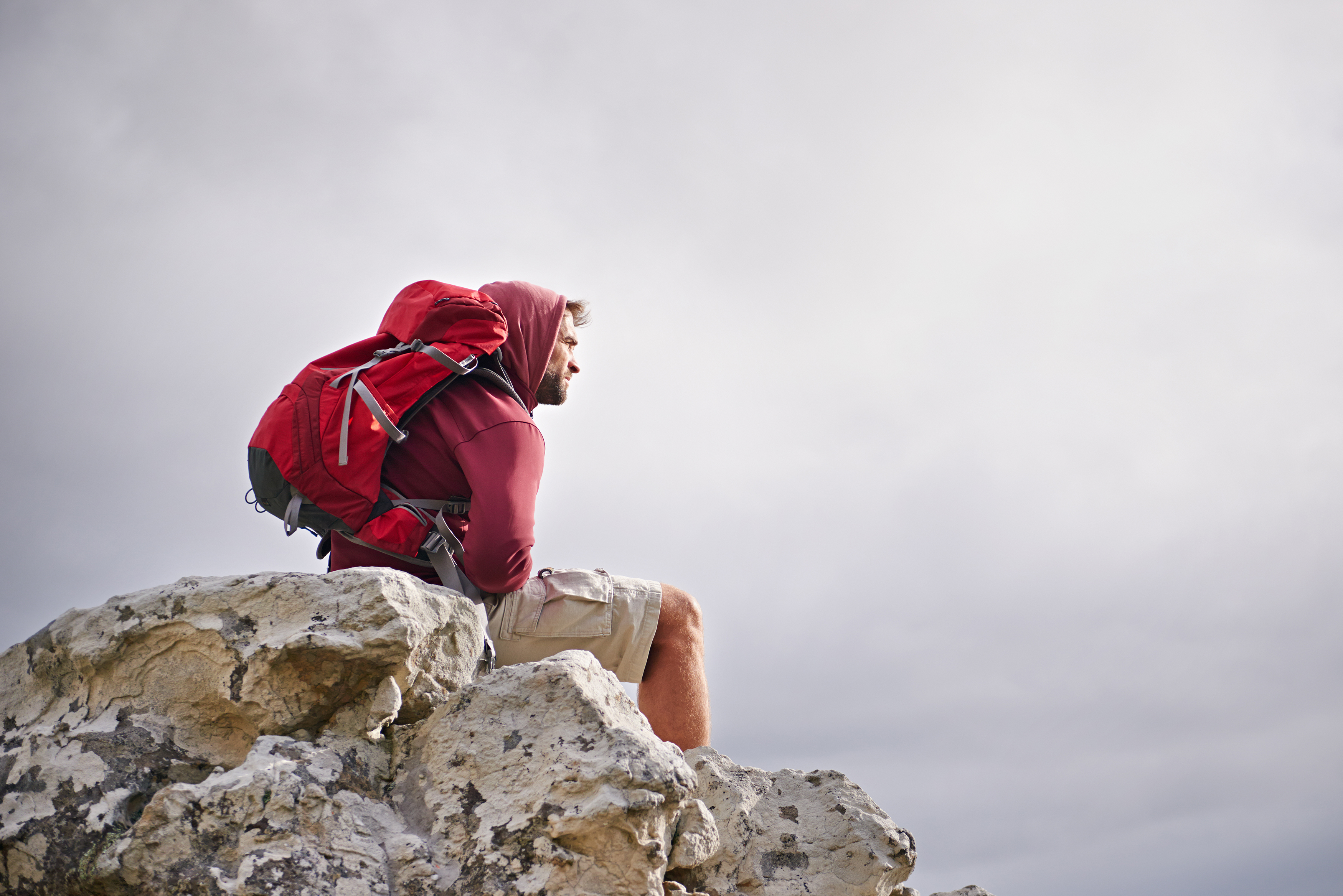 A man wearing a hooded jacket and a large red backpack sits on a rocky outcrop, facing to the right.