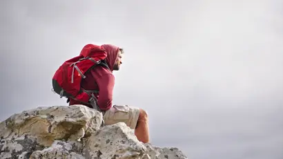 A man wearing a hooded jacket and a large red backpack sits on a rocky outcrop, facing to the right.