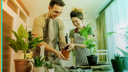 Two people wearing aprons stand at a table potting a houseplant together.